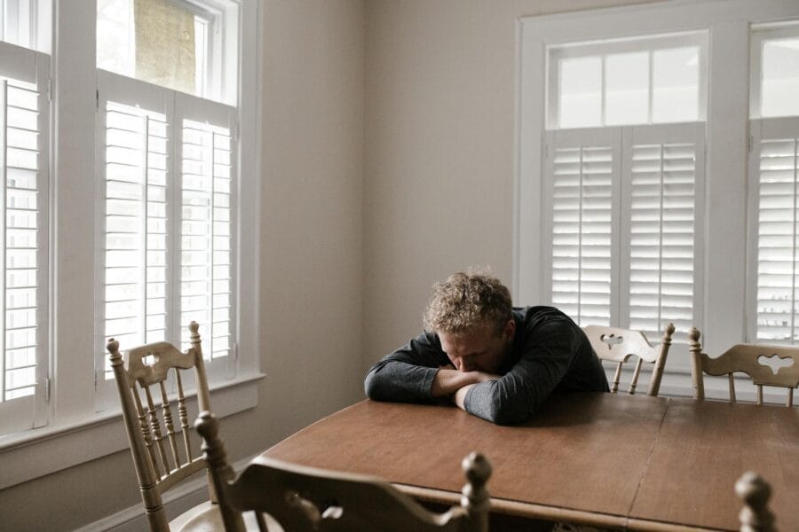 An adult man with emotions of sadness and anxiety sitting alone at a wooden table near windows.