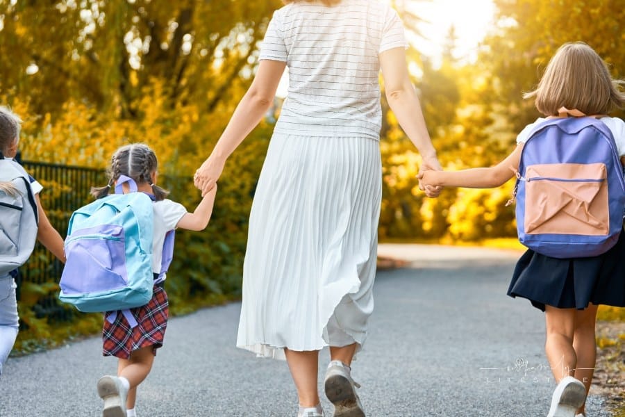mother wearing long white skirt walking her three daughters to school with backpacks on