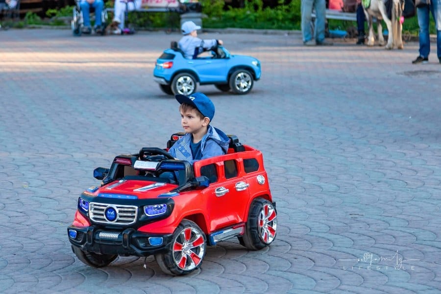 Two little boys ride on red and blue electric cars while walking with their parents in a city park with green trees, laughing