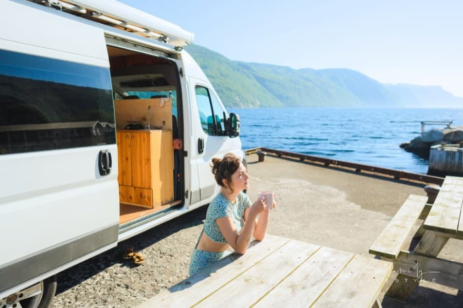 Woman sitting at the table and drinking coffee near camper van parked with view on fjord