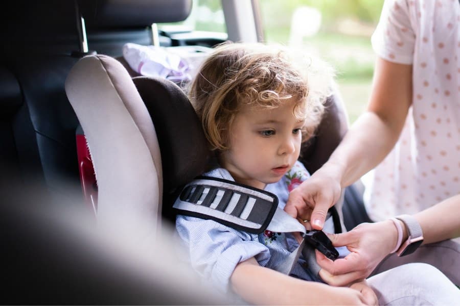 Mother putting small toddler daughter in car seat before trip, wearing face masks.