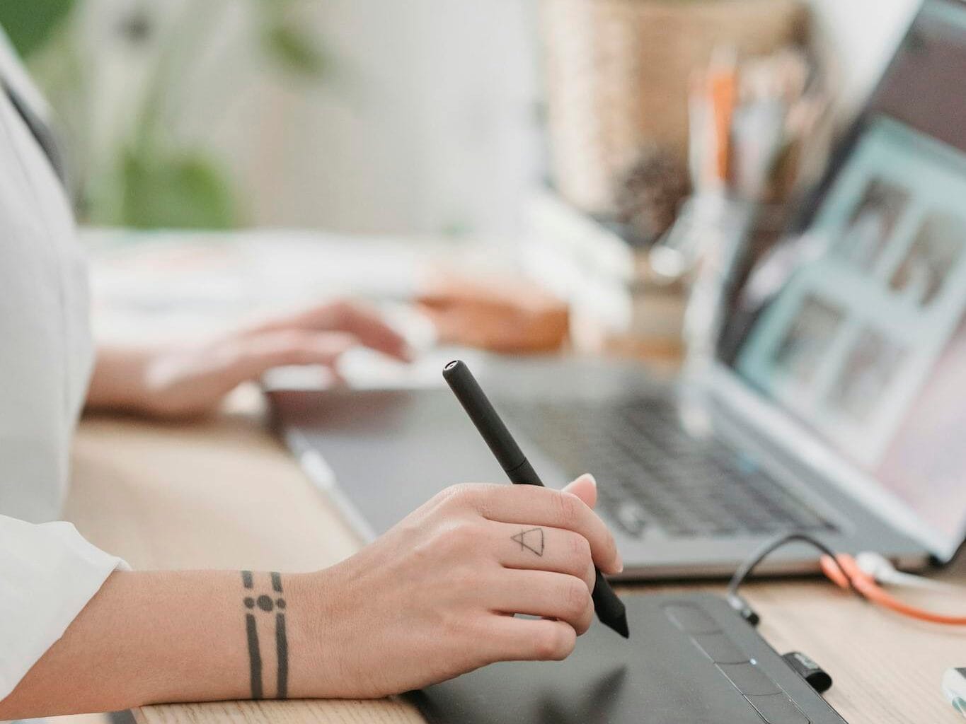 Crop anonymous female browsing netbook and working on graphic tablet with stylus while sitting at desk