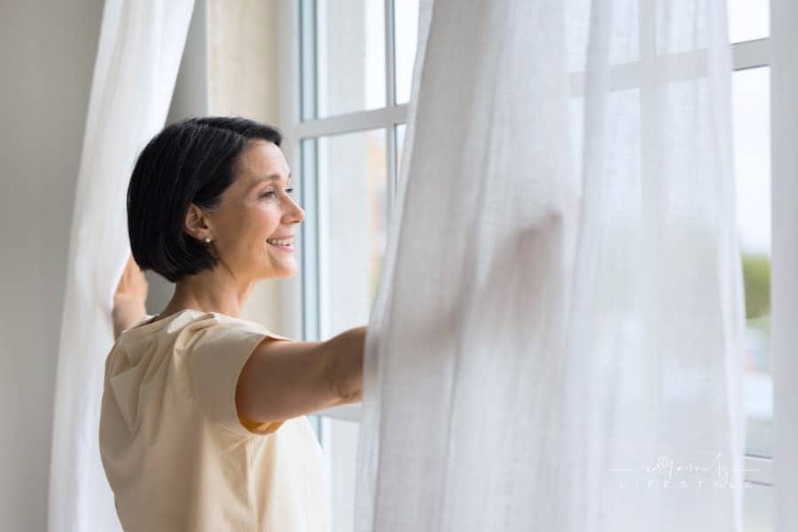 Happy mature homeowner woman looking out of big window
