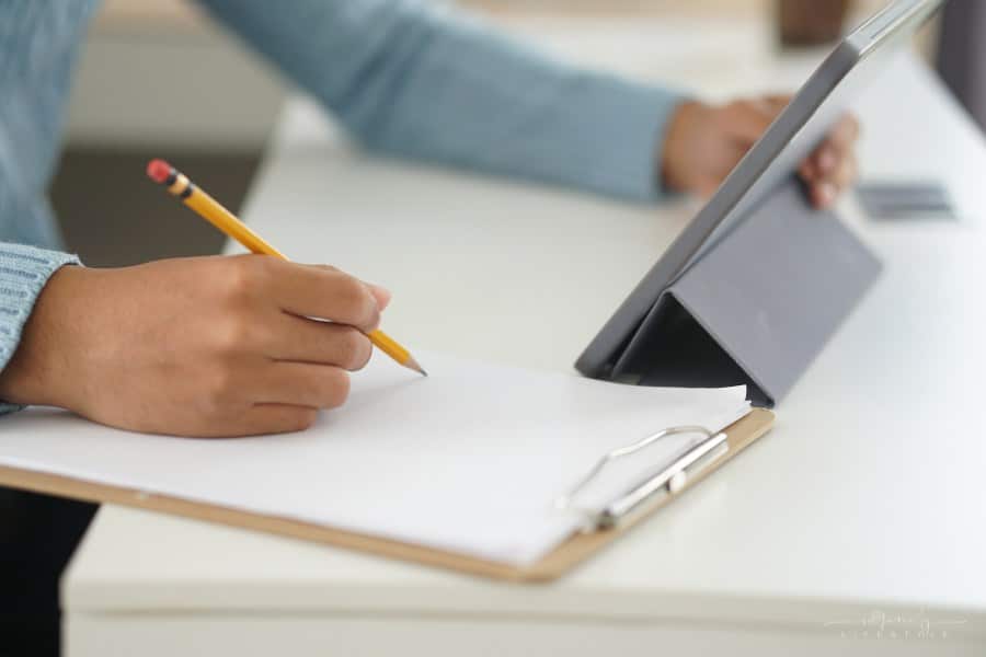male student writing on paper notebook while looking at digital tablet