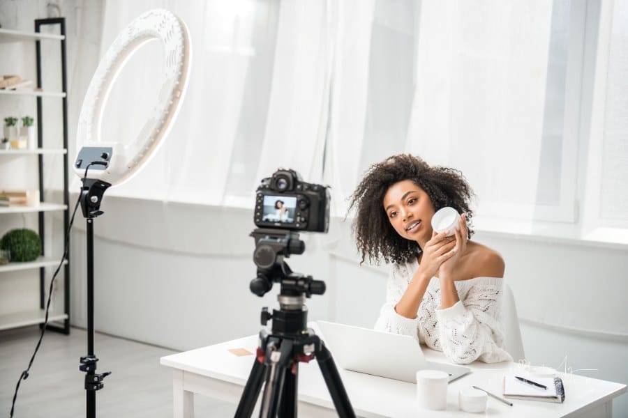 selective focus of attractive african american influencer in braces holding container with cosmetic cream near digital camera