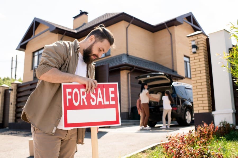 bearded man in casual shirt placing For sale sign against house to pay attention of buyers