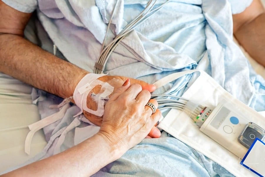 wife holding husband's hand as he lays in a hospital bed