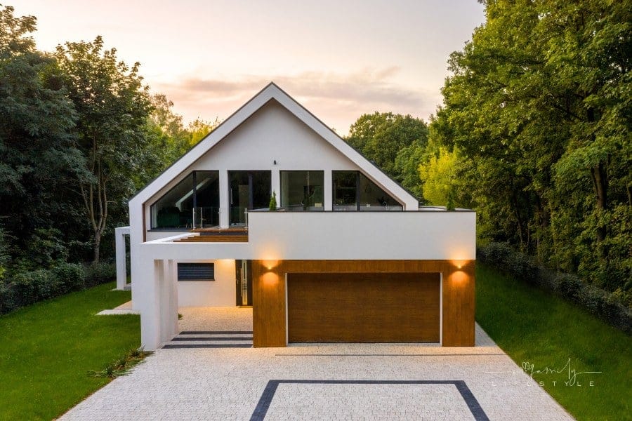 Elegant house with balcony, garage, cobblestone driveway, green lawn and lights on, exterior view during sunset