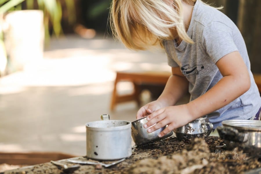 preschool boy playing in outdoor mud kitchen with pots and pans