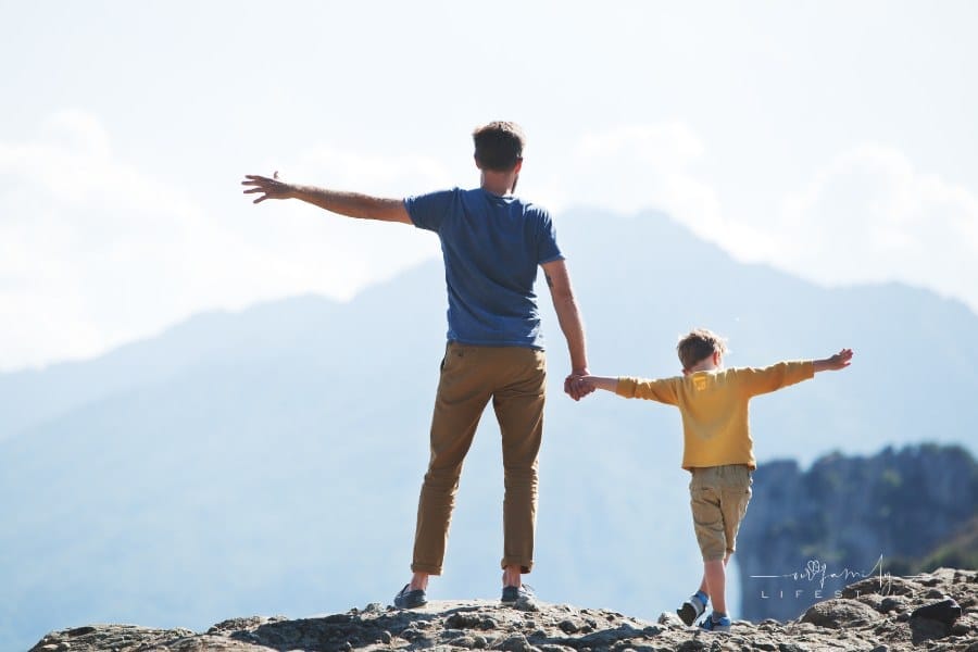 father and son holding hands and spreading arms wide on a mountain top