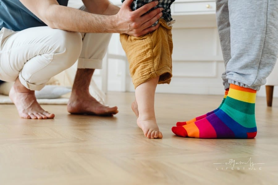 feet of LGBTQ couple Holding standing baby between them; one wearing rainbow pride socks