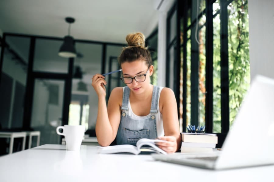 young woman wearing overalls reading a book while sitting in front of online learning class
