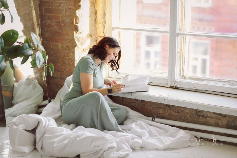 Young brunette woman relaxing at home alone reading favorite magazine while sitting on parquet floor on top of rolled-up bed