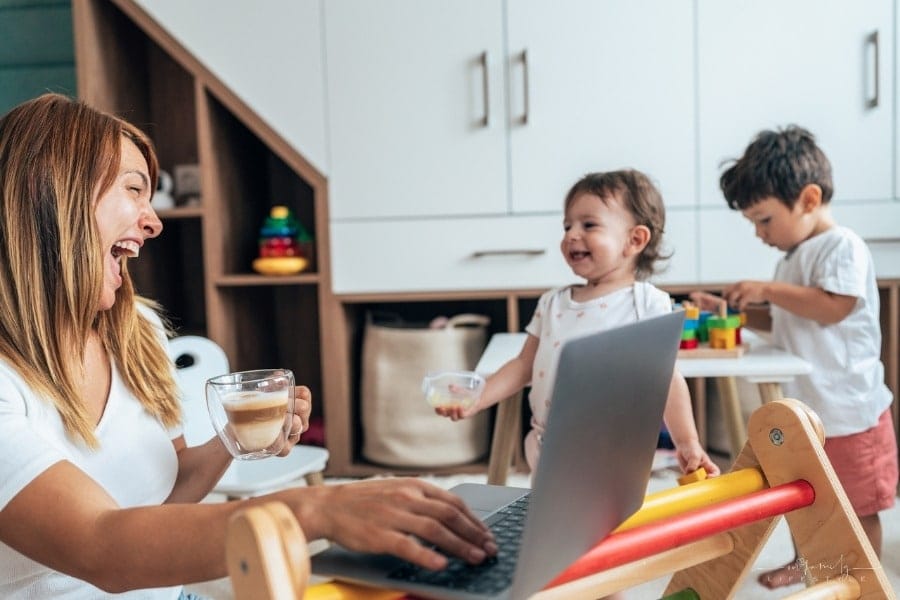 mom with wo small children working on laptop while they play nearby
