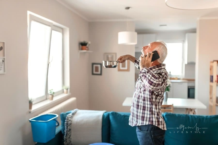 Angry Bearded Senior Man Talking on Smartphone Near Plastic Wash Bowl at Home in the Living Room Because of Roof Leaking