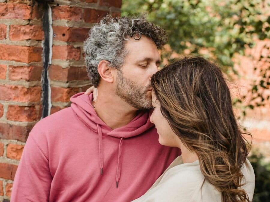 Romantic man in stylish clothes touching forehead of pregnant wife with closed eyes while resting together in green park near
