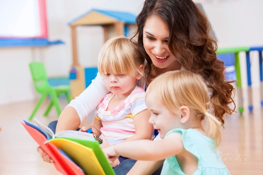 daycare teacher sitting on the floor and reading a colorful book to two preschool girls. The little girls are interested in t
