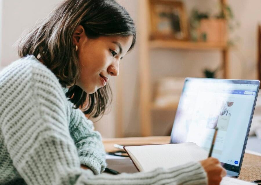 Side view of smart focused female writing in notebook while using netbook at table on blurred background