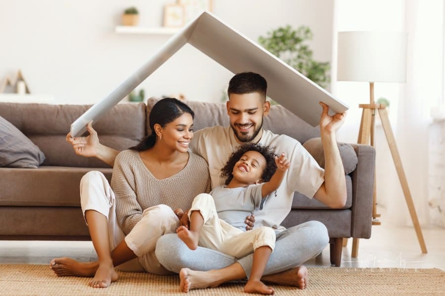 Delighted diverse parents and mixed race son sitting under roof and smiling near sofa in cozy living room at home