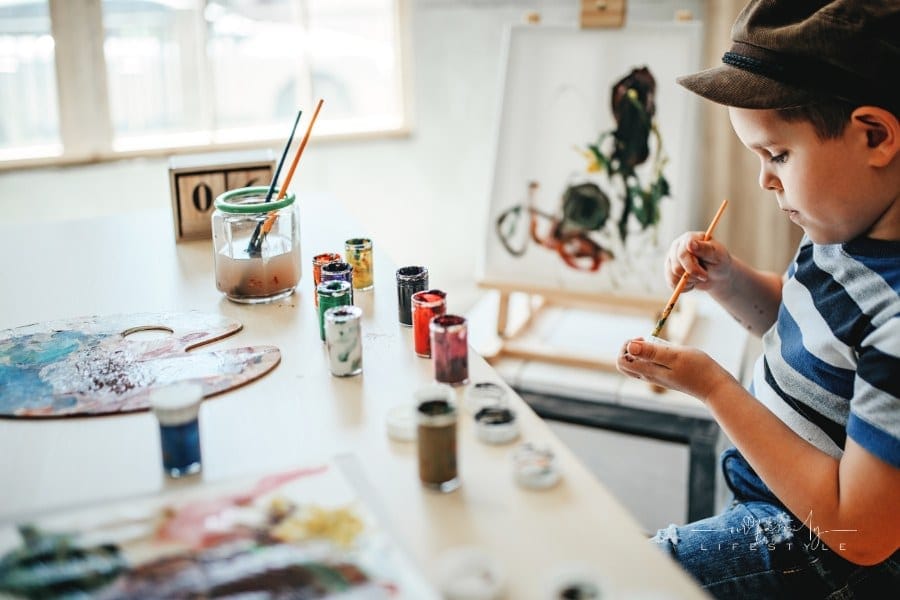 young boy sitting at table with paints, an easel, and a paintbrush in hand