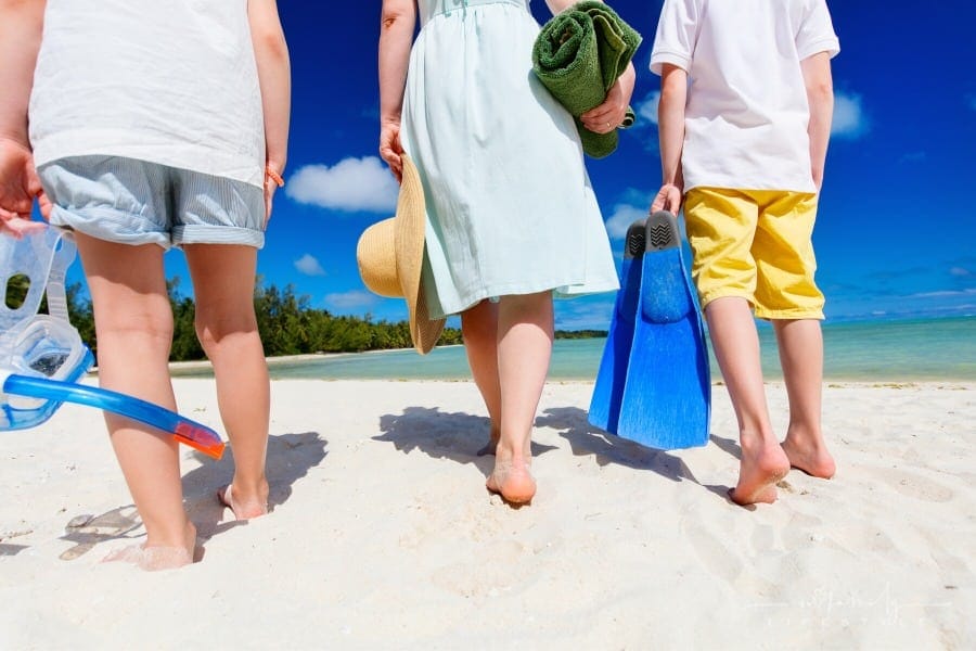 mother and kids walking on beach with snorkeling equipment, towel and sun hat