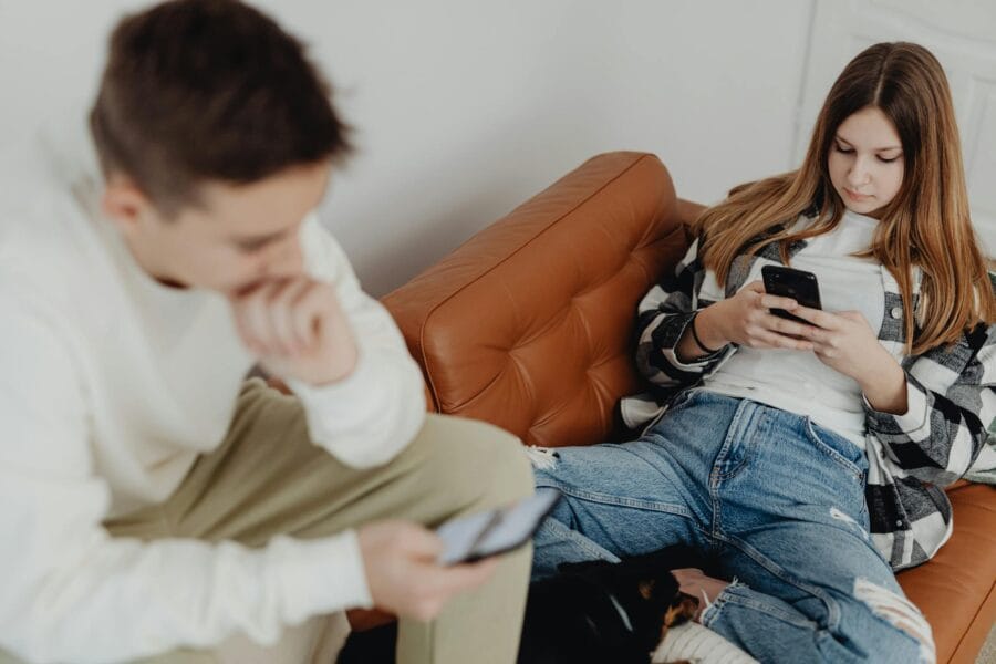 Two teenagers using smartphones while sitting comfortably on a leather couch indoors.
